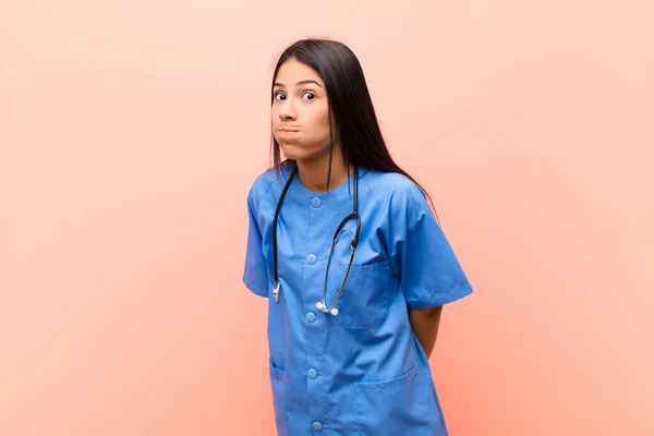 young latin nurse with a goofy, crazy, surprised expression, puffing cheeks, feeling stuffed, fat and full of food against pink wall