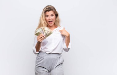 Genç, güzel, Latin bir kadın ağzı açık bir şekilde şaşkın ve şaşırmış görünüyor. Kendini dolar banknotlarıyla işaret ediyor.