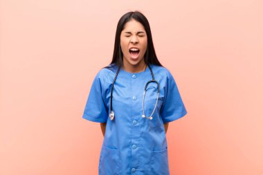 Genç Latin hemşire agresif bir şekilde bağırıyor, sinirli, sinirli, öfkeli ya da sinirli görünüyor, pembe duvara karşı hayır diye bağırıyor.