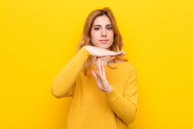 Genç, güzel sarışın kadın ciddi, sert, kızgın ve hoşnutsuz görünüyor, sarı duvara karşı zaman aşımı işareti yapıyor.