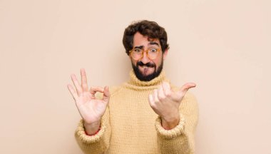 Havalı genç adam kendini mutlu, şaşkın, tatmin olmuş ve şaşırmış hissediyor, tamam gösteriyor ve jestler yapıyor, düz duvara gülümsüyor.