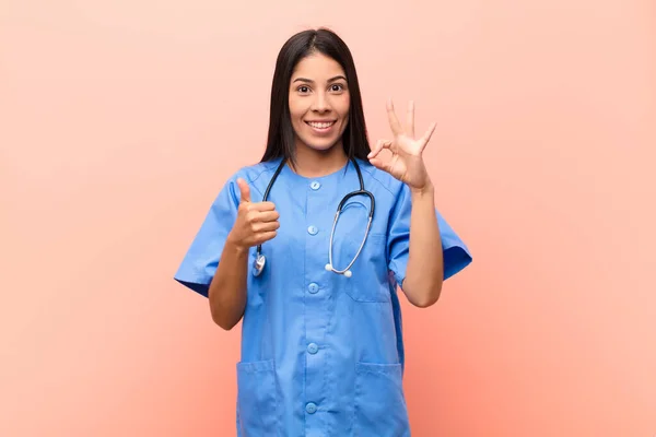 young latin nurse feeling happy, amazed, satisfied and surprised, showing okay and thumbs up gestures, smiling against pink wall