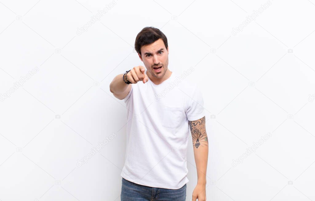 young handsome man pointing at camera with an angry aggressive expression looking like a furious, crazy boss against white wall