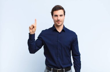 Genç yakışıklı adam neşeyle ve mutlulukla gülümsüyor, tek eliyle yukarıyı işaret ederek mavi duvara uzayı kopyalıyor.