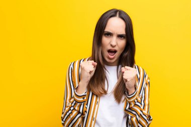 Genç ve güzel bir kadın sinirli sinirli, sinirli bakışlar ve yumruklarla bağırıyor. Turuncu duvara karşı öfkeli hissediyor.