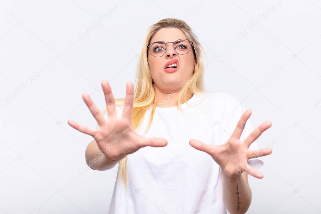 young pretty blonde woman feeling terrified, backing off and screaming in horror and panic, reacting to a nightmare against white wall