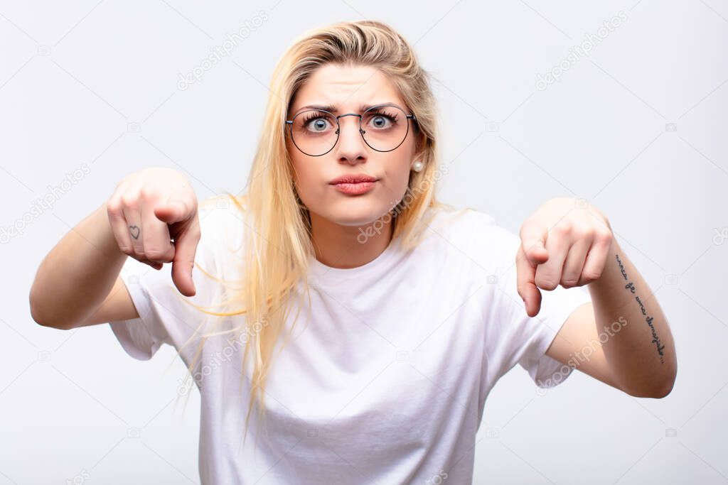 young pretty blonde woman pointing forward at camera with both fingers and angry expression, telling you to do your duty against white wall