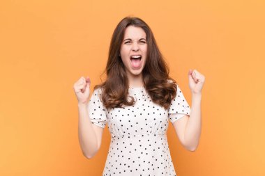 Genç, güzel bir kadın mutlu, şaşırmış ve gururlu hissediyor, bağırıyor ve başarıyı kutluyor turuncu duvarda kocaman bir gülümsemeyle.