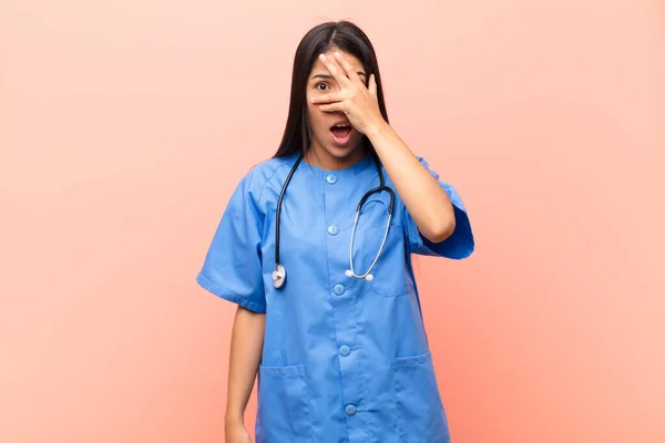 若いラテン語の看護師はショックを受け恐怖を感じました手で顔を覆いピンクの壁に指を覗かせました — ストック写真