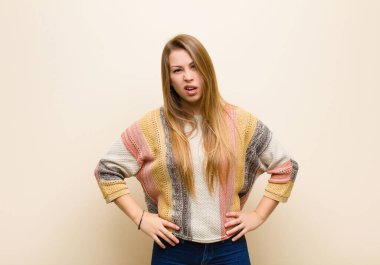 young blonde woman feeling puzzled and confused, with a dumb, stunned expression looking at something unexpected against beige wall