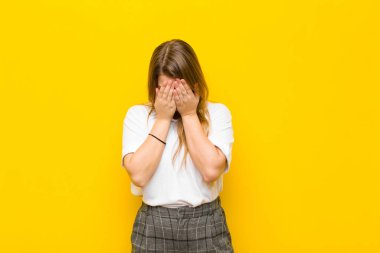 Genç sarışın kadın üzgün, hayal kırıklığına uğramış, gergin ve depresif, yüzünü iki eliyle kapatmış, turuncu duvara karşı ağlıyor.