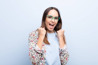 Genç ve güzel bir kadın zaferle bağırıyor, gülüyor, mutlu ve heyecanlı hissediyor mavi duvara karşı başarıyı kutlarken.