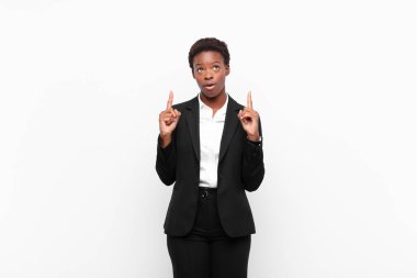 Genç, güzel, siyah bir kadın, beyaz duvara karşı şaşkın ve şaşkın bir bakışla yukarıyı işaret ediyor.