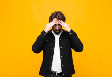 Genç çılgın yakışıklı adam stresli ve sinirli görünüyor, baskı altında çalışıyor, başı ağrıyor ve turuncu duvara karşı sorunları var.