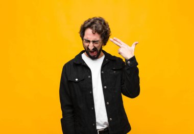 Genç ve yakışıklı adam mutsuz ve stresli görünüyor, intihar jesti elle silah işareti yapıyor, kafasını turuncu duvara doğrultuyor.