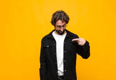 Genç, çılgın, yakışıklı, neşeli ve gelişigüzel gülümseyen, aşağıya bakan ve göğsünü turuncu duvara gösteren bir adam.