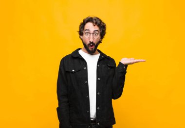 Genç çılgın yakışıklı adam şaşırmış ve şok olmuş görünüyor, çenesi düşmüş, bir objeyi tutarken elini turuncu duvara karşı açık tutuyor.