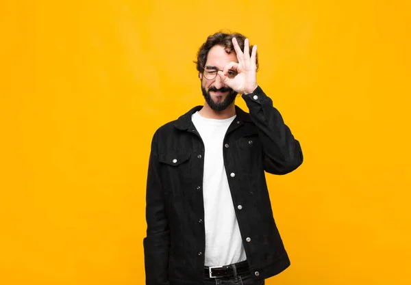 young crazy handsome man smiling happily with funny face, joking and looking through peephole, spying on secrets against orange wall