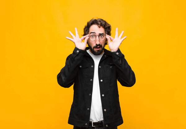 young crazy handsome man feeling shocked, amazed and surprised, holding glasses with astonished, disbelieving look against orange wall