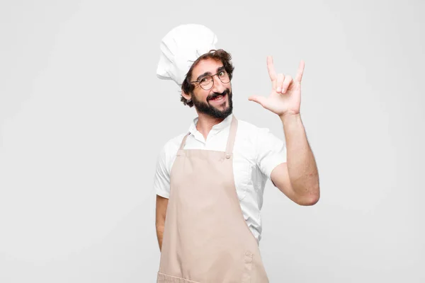 young crazy chef feeling happy, fun, confident, positive and rebellious, making rock or heavy metal sign with hand against white wall