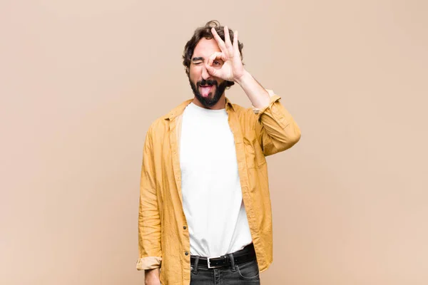 young crazy cool man smiling happily with funny face, joking and looking through peephole, spying on secrets against flat wall