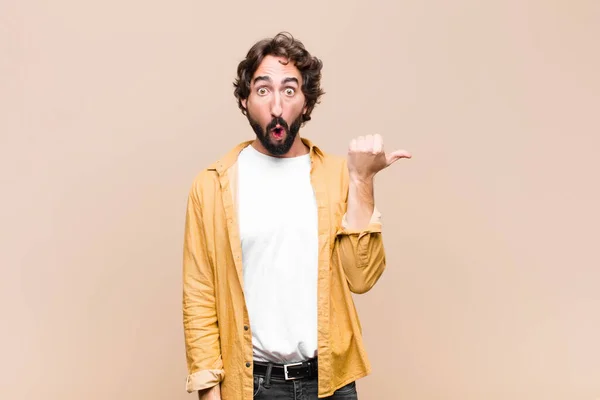 young crazy cool man looking astonished in disbelief, pointing at object on the side and saying wow, unbelievable against flat wall