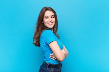 Genç, güzel bir kadın neşeyle gülümsüyor, mutlu, tatmin olmuş ve rahatlamış hissediyor, kollarını kavuşturup mavi duvara bakıyor.