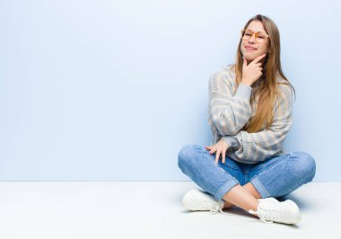 Genç sarışın kadın ciddi, düşünceli ve güvensiz görünüyor, bir kolu çapraz ve çene üzerinde, yerde oturan seçenekleri tartıyor