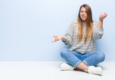 Genç sarışın kadın, aptal, deli, kafası karışık, şaşkın bir ifadeyle omuz silkiyor, yerde oturmaktan rahatsız ve bihaber hissediyor.