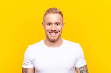 Genç sarışın yakışıklı adam kameraya gülümsüyor ve mutlu, kendinden emin, tatmin olmuş bir yüz ifadesi var. sarı duvara yaslan