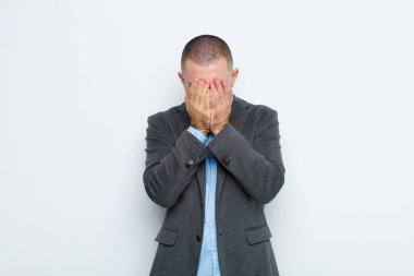 Genç işadamı üzgün, hayal kırıklığına uğramış, gergin ve depresif hissediyor, iki eliyle yüzünü kapatıyor, düz duvara karşı ağlıyor.