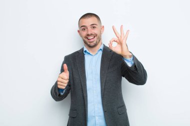 Genç işadamı kendini mutlu, şaşkın, tatmin olmuş ve şaşırmış hissediyor, tamam gösteriyor ve jestler yapıyor, düz duvara gülümsüyor