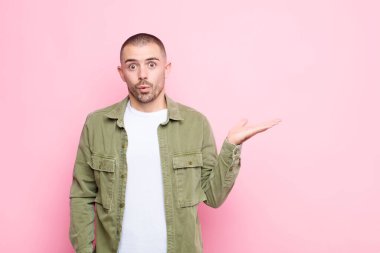 Genç yakışıklı adam şaşırmış ve şoka uğramış görünüyor, çenesi düşmüş ve düz duvara karşı eli açık bir nesne tutuyor.