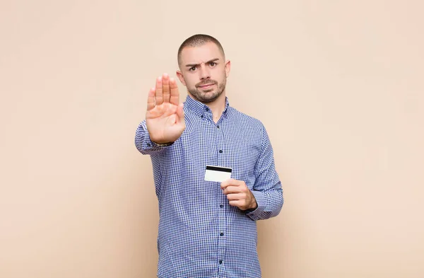 年轻英俊的男人看上去严肃 不高兴 用信用卡摆出摊开手掌的停止姿势 — 图库照片