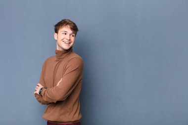 Genç sarışın adam neşeyle gülümsüyor, mutlu, tatmin olmuş ve rahatlamış hissediyor, kollarını kavuşturup düz duvara bakıyor.