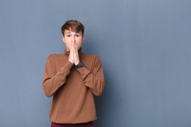 Genç sarışın adam endişeli, üzgün ve korkmuş hissediyor, elleriyle ağzını kapatıyor, endişeli görünüyor ve düz duvara karşı izole edilmiş.