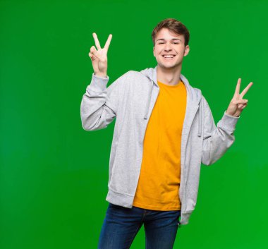 Genç sarışın adam gülümsüyor ve mutlu görünüyor, arkadaş canlısı ve memnun, işaret ediyor zafer ya da barış iki eliyle kroma anahtar duvara karşı