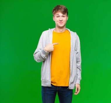 Genç sarışın adam gururlu, kendinden emin ve mutlu görünüyor, gülümsüyor ve kendini işaret ediyor ya da kroma tuşuna karşı bir numara işareti yapıyor.