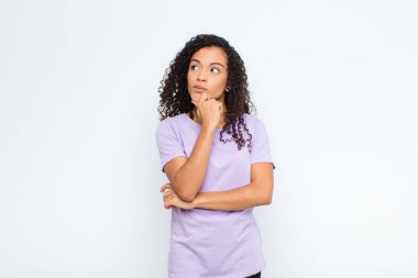 Genç siyahi kadın düşünüyor, kuşkulu ve şaşkın hissediyor, farklı seçeneklerle, beyaz duvara karşı hangi kararı vereceğini merak ediyor.