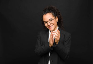 young black woman feeling happy and successful, smiling and clapping hands, saying congratulations with an applause against black wall clipart