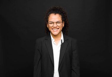 Genç siyah kadın mutlu ve şapşal görünüyor. Geniş, eğlenceli, kaçık bir gülüşü ve kara duvarın karşısında gözleri açık.