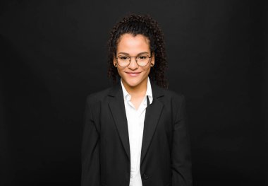 Genç siyahi kadın pozitif ve kendinden emin gülümsüyor. Tatmin olmuş, arkadaş canlısı ve siyah duvara karşı mutlu görünüyor.