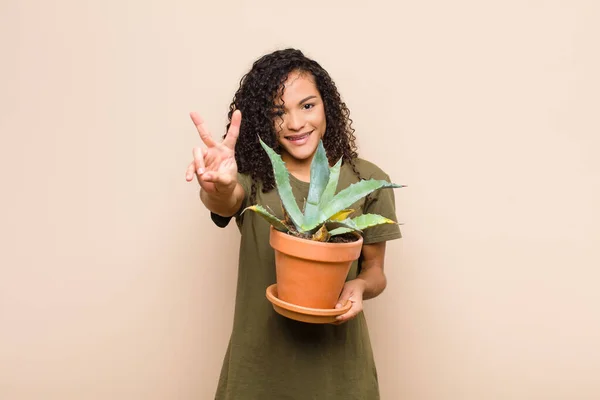 young black woman smiling and looking happy, carefree and positive, gesturing victory or peace with one hand holding a cactus