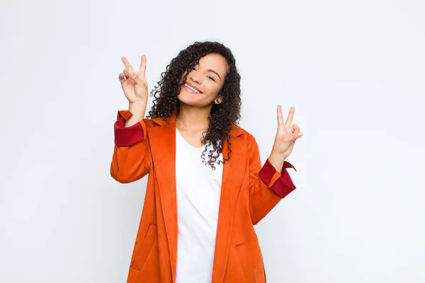 young black woman smiling and looking happy, friendly and satisfied, gesturing victory or peace with both hands against white wall