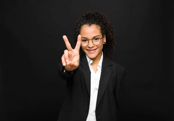 young black woman smiling and looking happy, carefree and positive, gesturing victory or peace with one hand against black wall