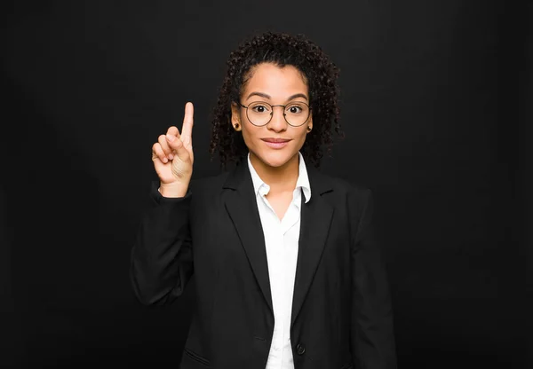 young black woman smiling cheerfully and happily, pointing upwards with one hand to copy space against black wall