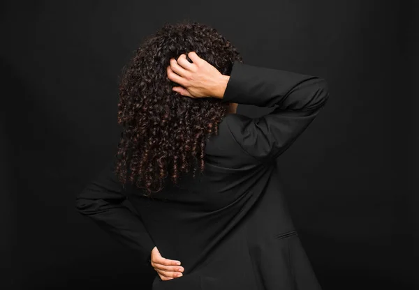 young black woman thinking or doubting, scratching head, feeling puzzled and confused, back or rear view against black wall