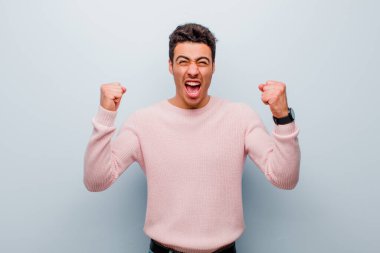 Genç Arap adam öfkeli bir ifadeyle ya da gri duvara karşı başarıyı kutlayan yumruklarla agresif bir şekilde bağırıyor.
