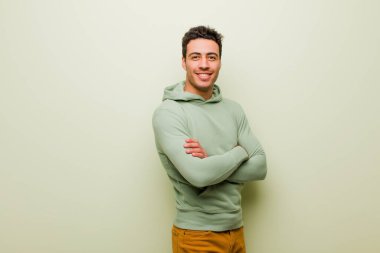 Genç Arap adam kameraya çapraz kollarla gülümsüyor ve mutlu, kendinden emin, tatmin olmuş bir ifade düz duvara karşı yan bakış.