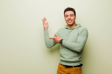 Genç Arap adam gururla ve güvenle gülümsüyor, mutlu ve tatmin olmuş hissediyor ve düz duvara karşı fotokopi alanında bir konsept gösteriyor.